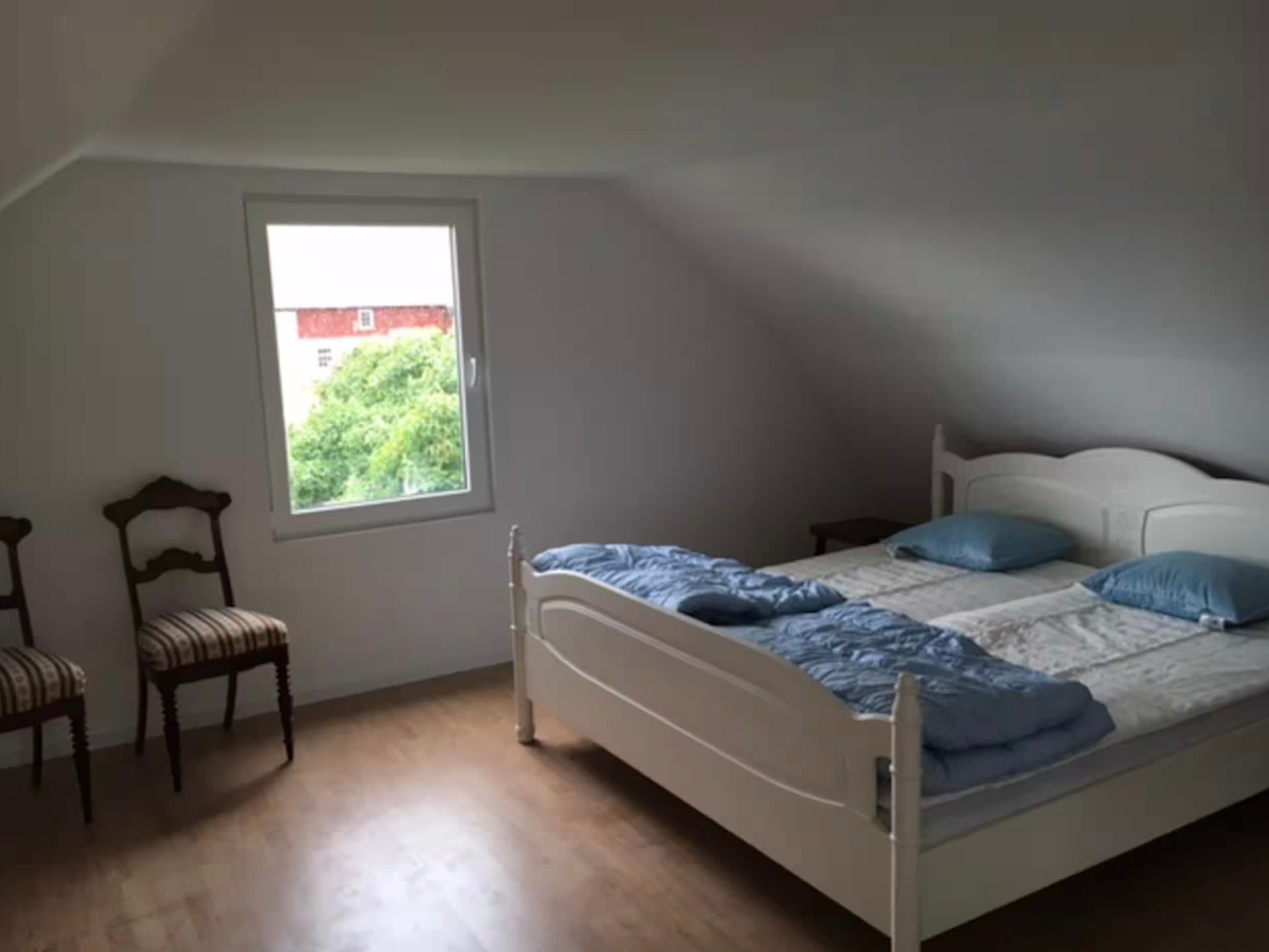Luftig sovrum med stora fönster och naturligt ljus. Bekvämt dubbelbed med blåa kuddar, klassiska trästolar och vyer över den gröna omgivningen.